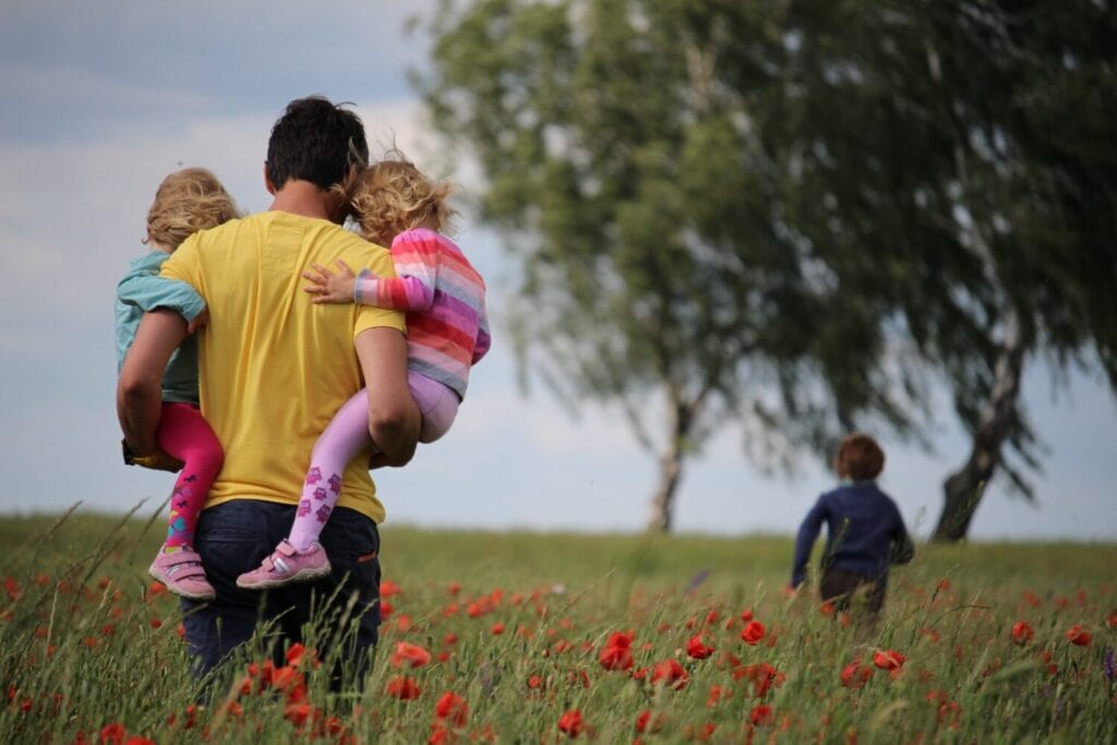 Mann trägt zwei Kinder über die Wiese und Junge rennt voraus