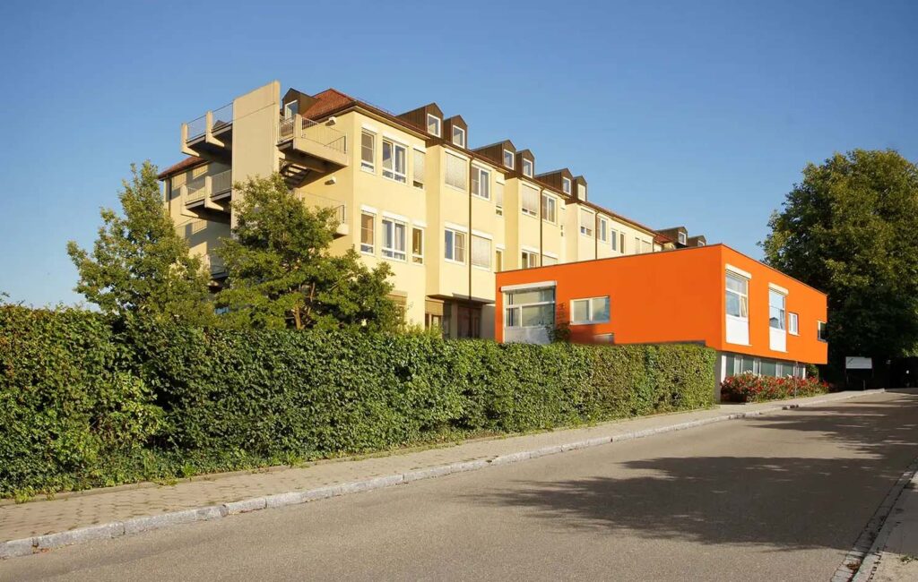 Modernes Gebaeude mit gelber Fassade und orangenem Anbau, umgeben von Gruen.