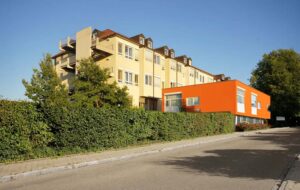 Modernes Gebaeude mit gelber Fassade und orangenem Anbau, umgeben von Gruen.