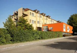 Modernes Gebäude mit gelber Fassade und orangenem Anbau, umgeben von Grün.