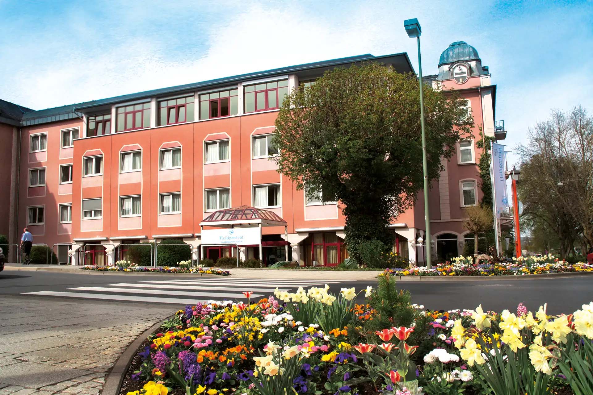 Rosa Gebäude mit Blumenbeeten im Vordergrund und blühenden Bäumen.