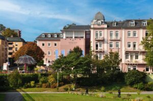 Rosa Klinikgebaeude mit Gartenanlagen und Baeumen vor blauem Himmel.
