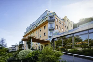 Modernes Klinikgebäude mit Balkon und Glasfassade, umgeben von Grün.