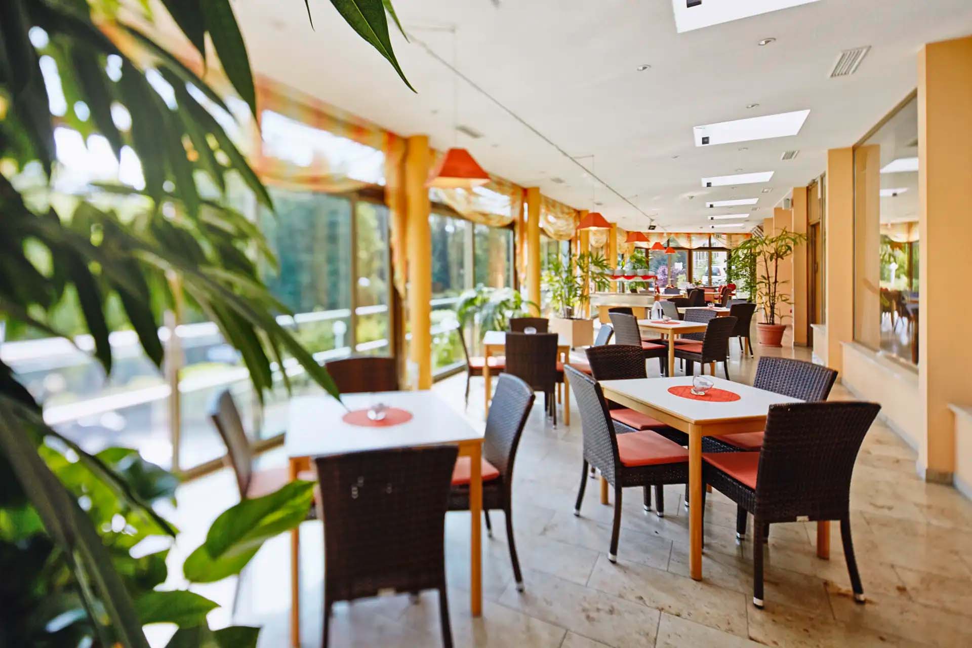 Heller Speisesaal mit großen Fenstern, Pflanzen und gemütlichen Sitzbereichen.