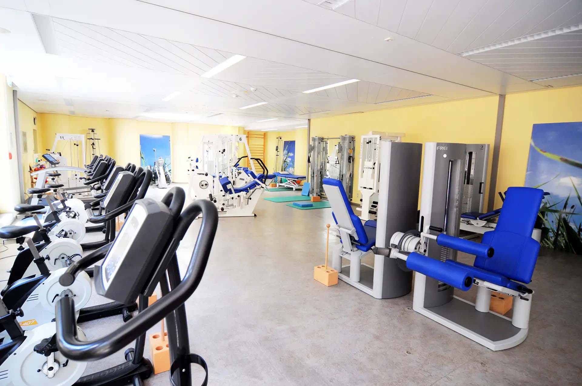 Fitnessraum mit verschiedenen Trainingsgeraeten und hellgelben Waenden.