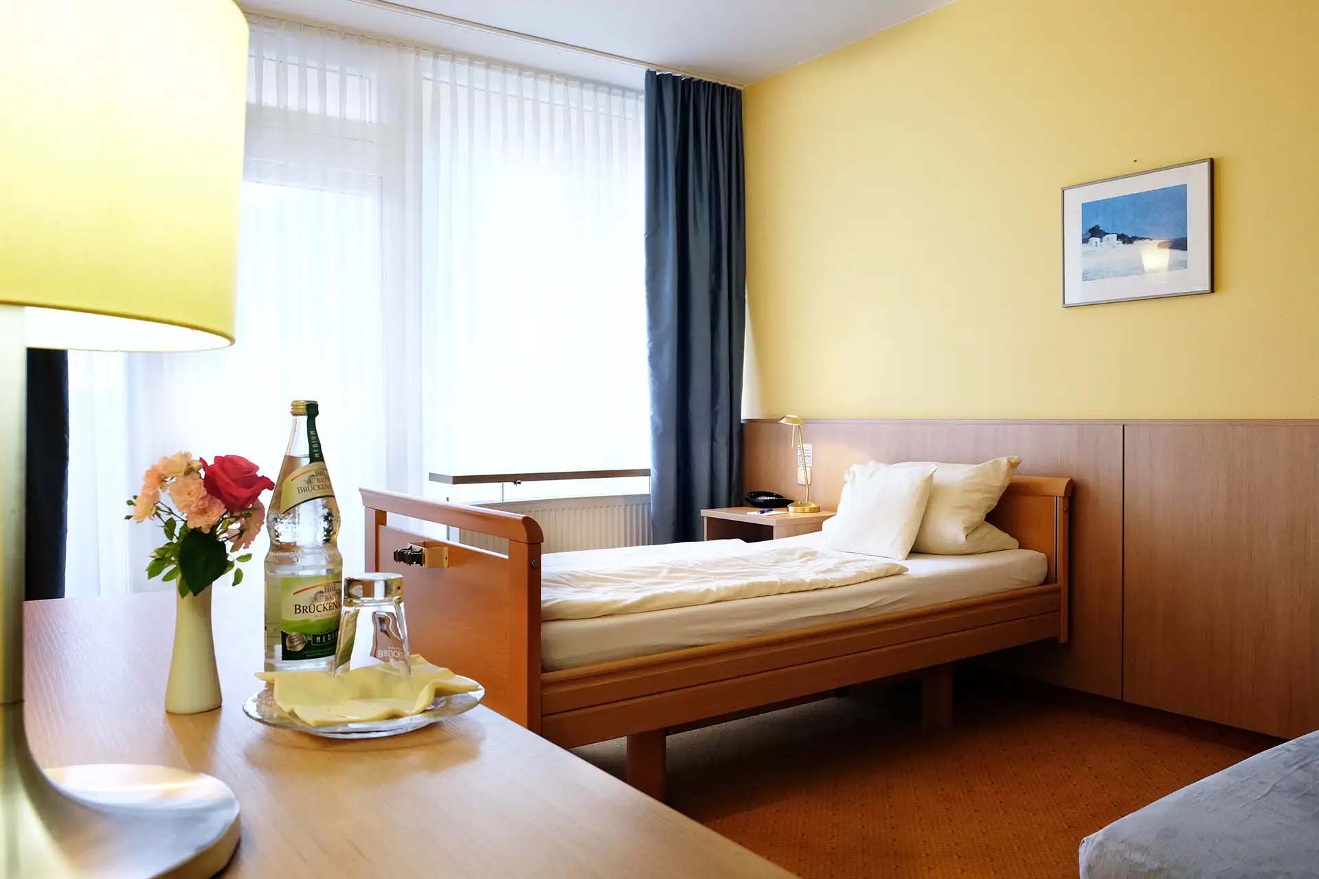 Helles Einzelzimmer mit Bett, Schreibtisch und Blumen in einer Klinik.