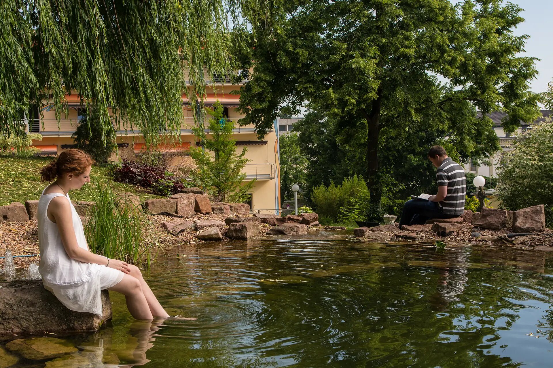 Menschen entspannen an einem Teich im Garten einer Psychosomatischen Klinik.