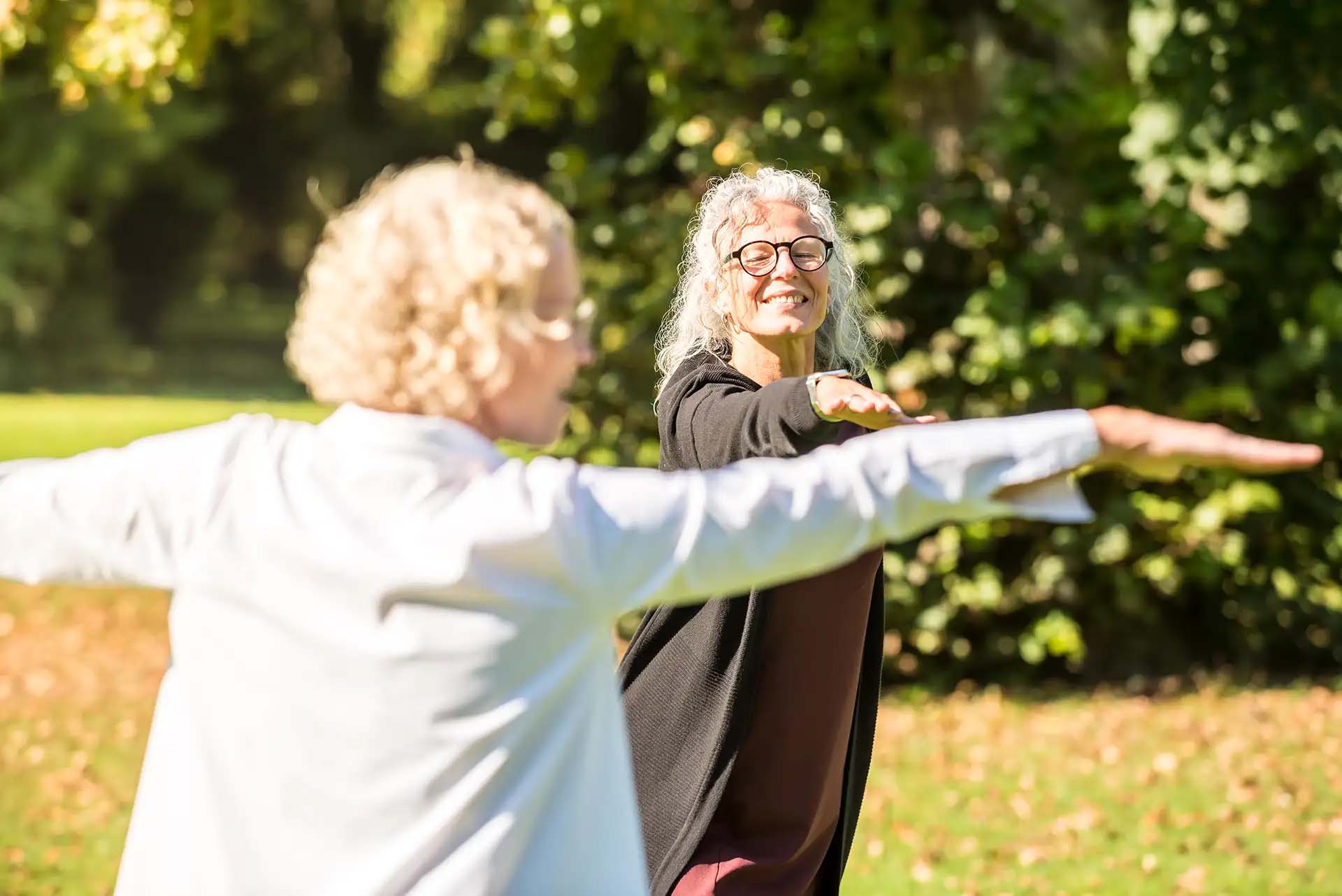 Zwei Frauen machen zusammen Yoga-Uebungen im Freien und laecheln dabei.