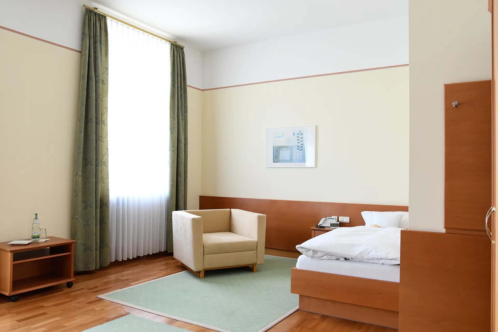 Helles Zimmer mit Bett, Sessel, grünem Teppich und Vorhängen.