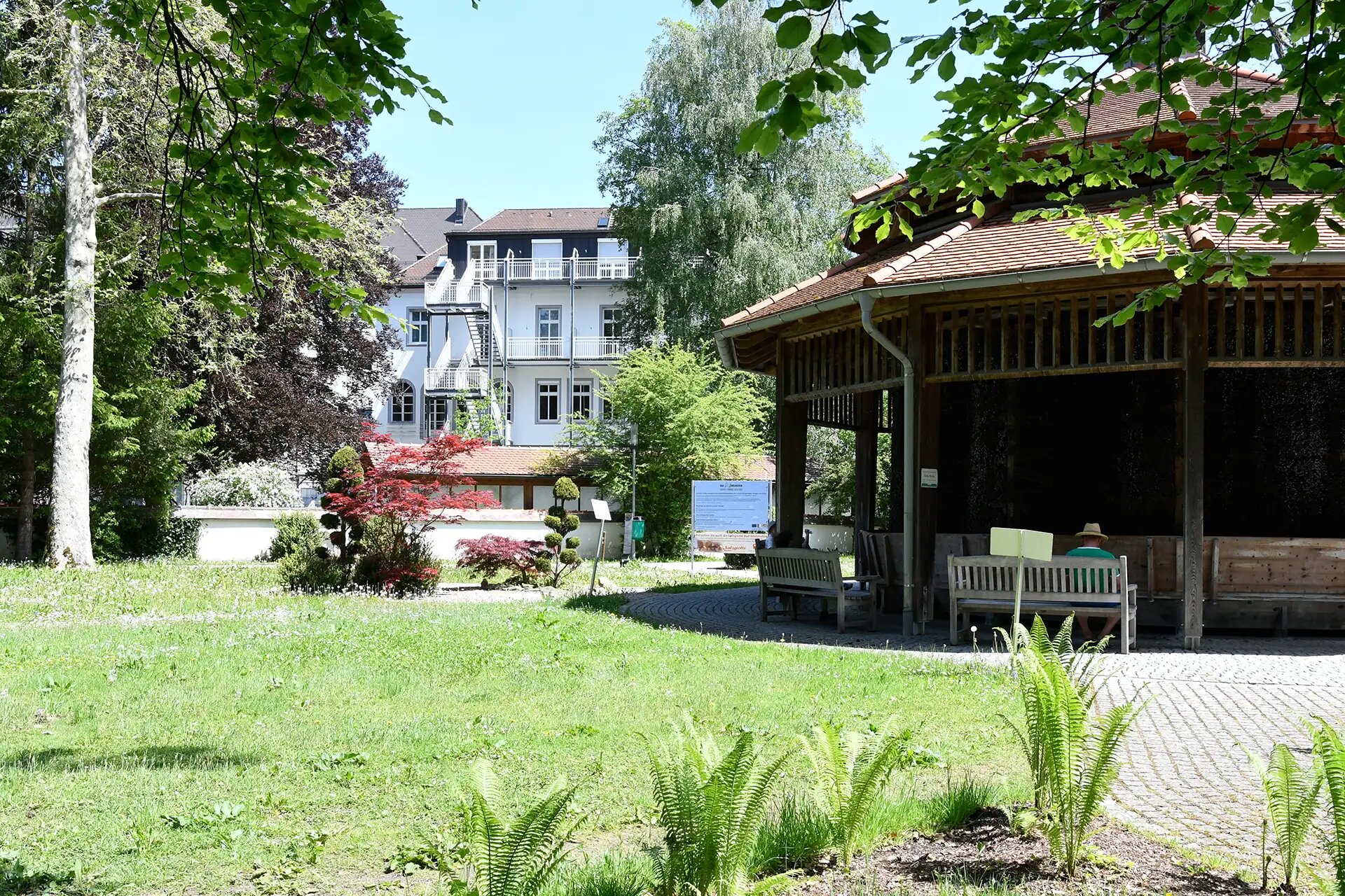 Garten mit Pavillon, Sitzbaenken, Blumen und Gebaeuden im Hintergrund.
