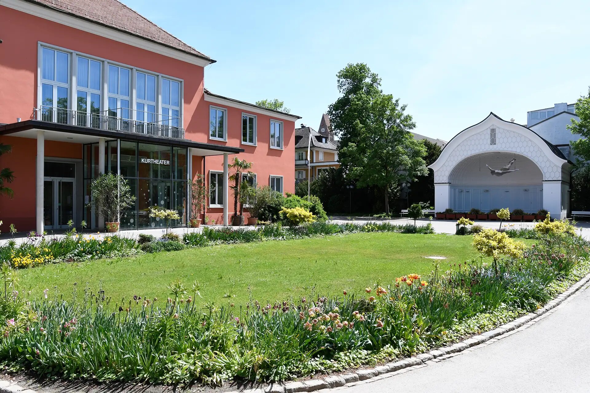 Kurtheater mit gepflegtem Garten und Springbrunnen im Sonnenschein.