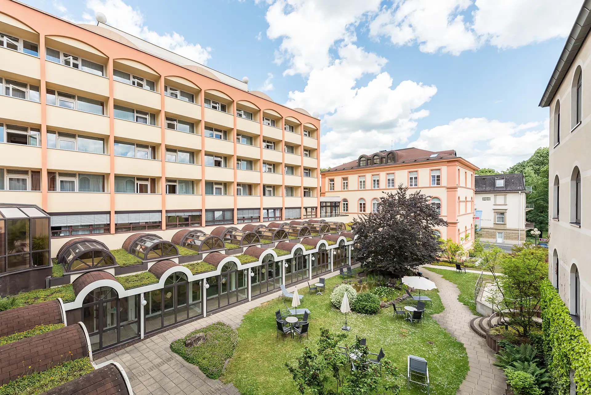 Hofansicht eines Gebaeudes mit Garten und Sitzgelegenheiten bei schoenem Wetter.