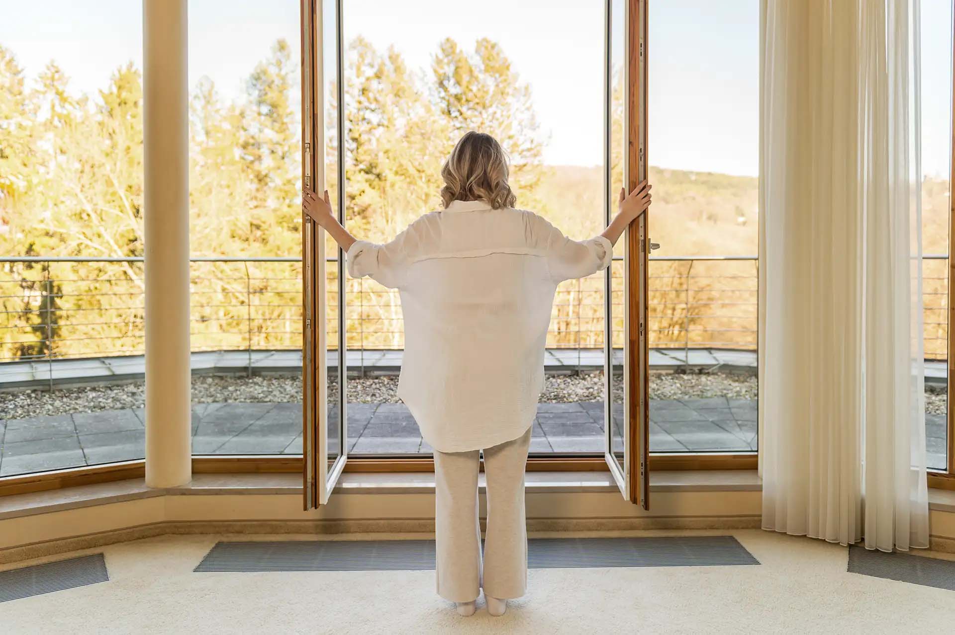 Frau öffnet Balkonfenster und blickt auf herbstliche Landschaft.