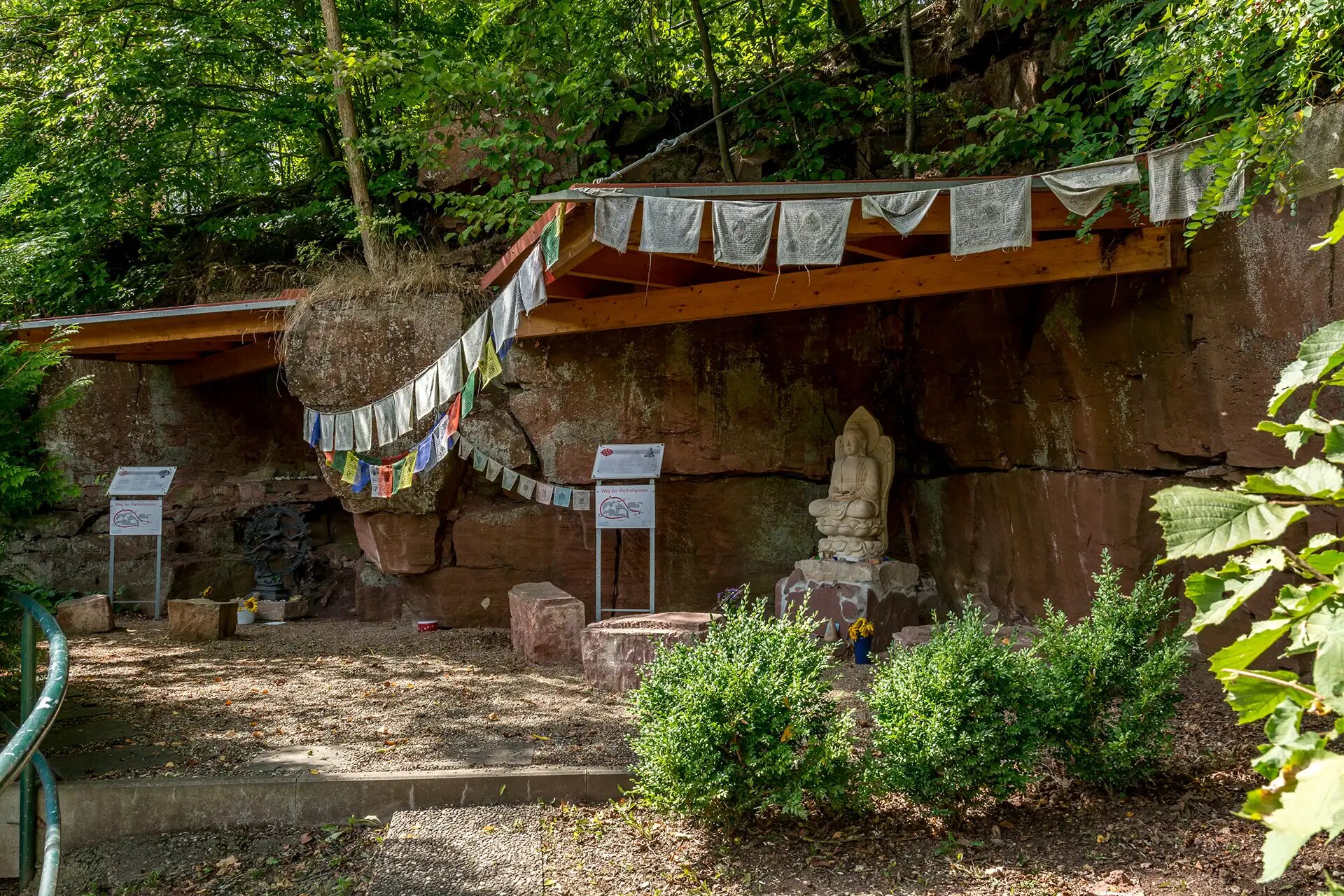 Buddahstatue vor einer Sandsteinmauer mit tibetischen Fahnen im Wald.