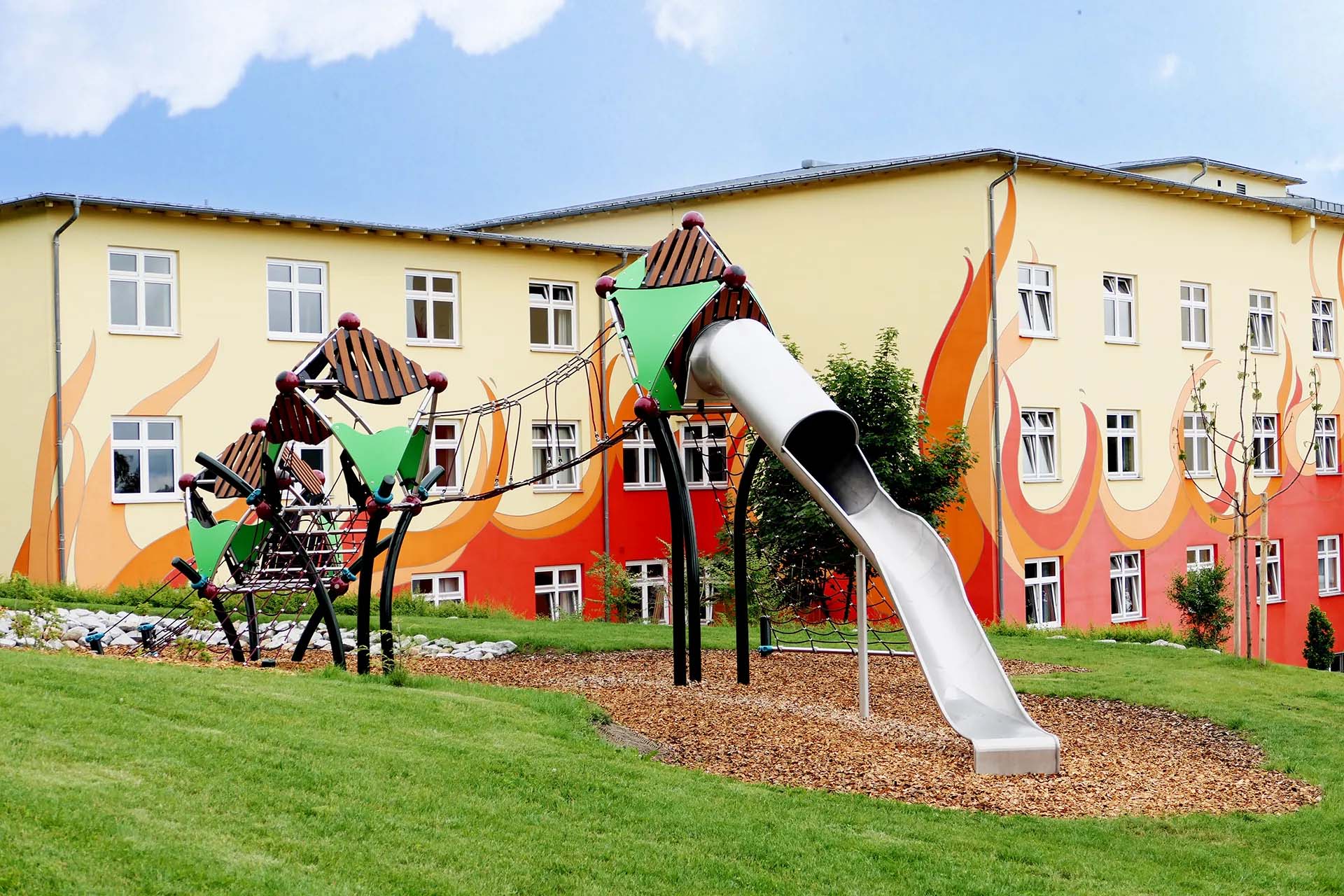 Bunter Spielplatz mit Rutsche vor einem farbenfrohen Gebäude mit Wandmalerei.