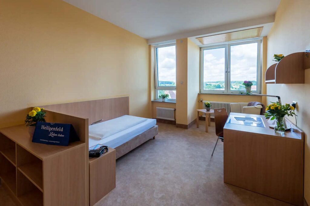 Geraeumiges Einzelzimmer mit Bett, Schreibtisch und großem Fenster mit Aussicht.