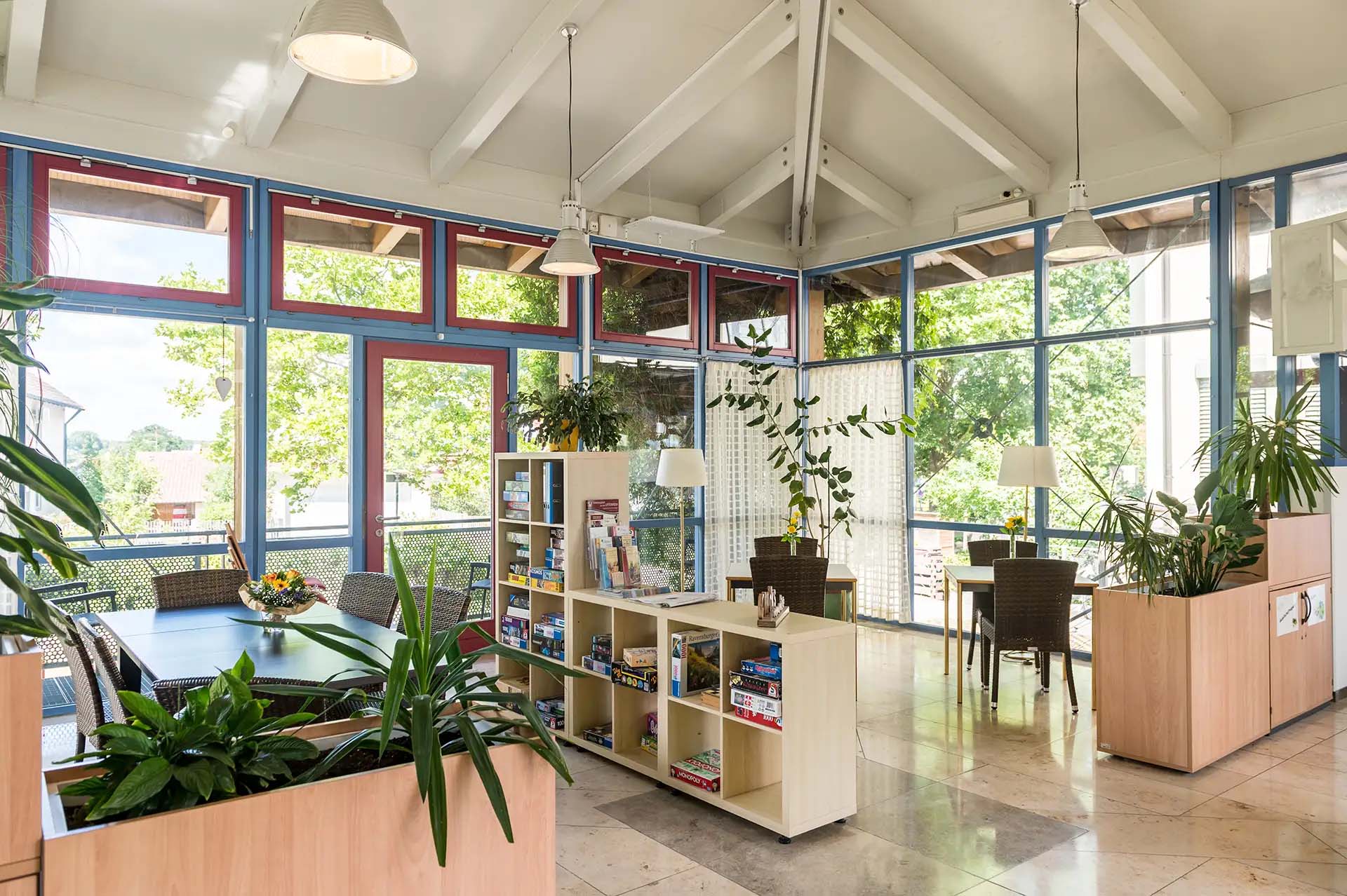 Helle Gemeinschaftslounge mit Pflanzen, Spielebibliothek und großen Fenstern.
