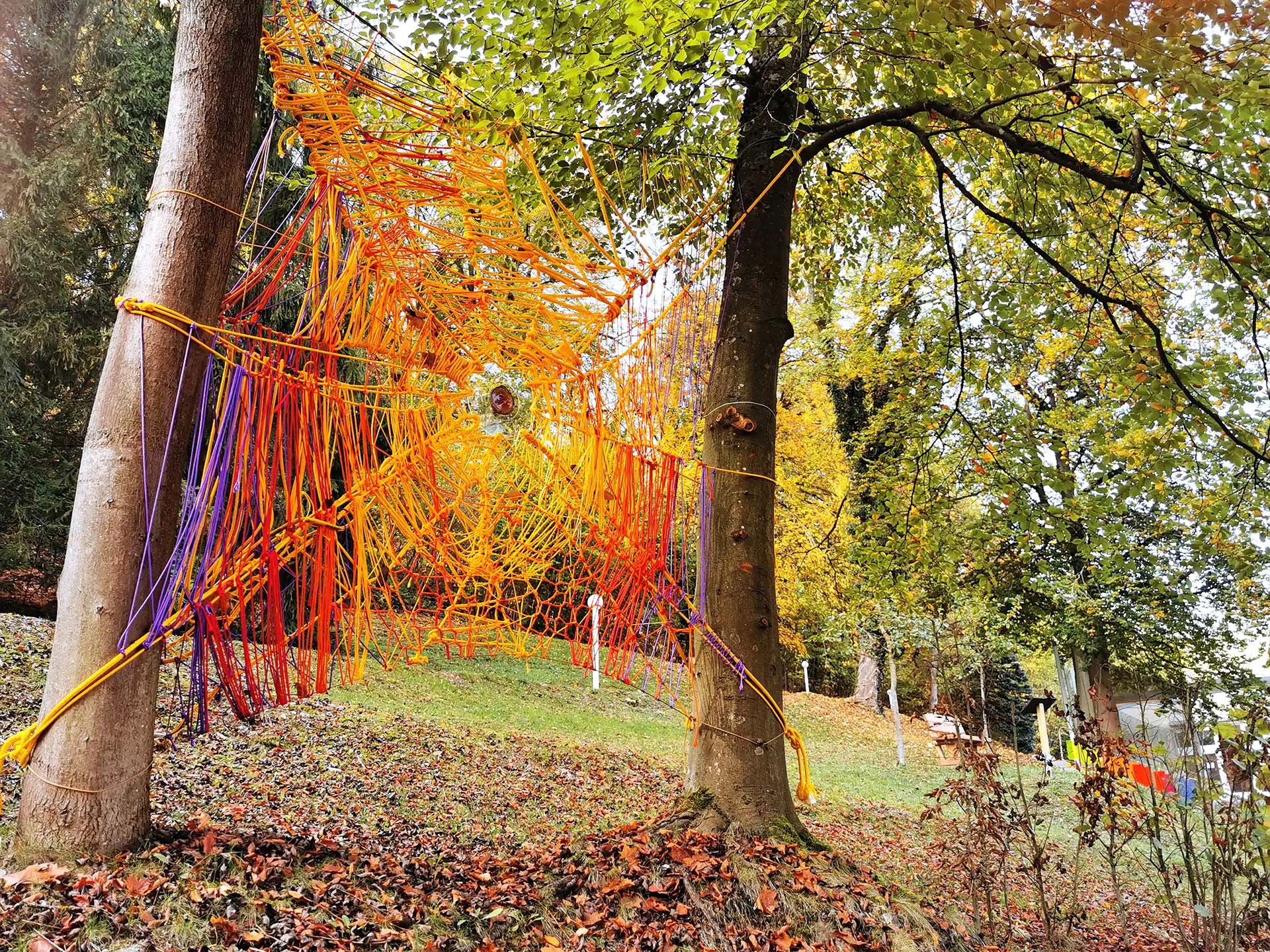 Farbenfrohes Netzkunstwerk aus Baendern zwischen Baeumen im herbstlichen Wald.