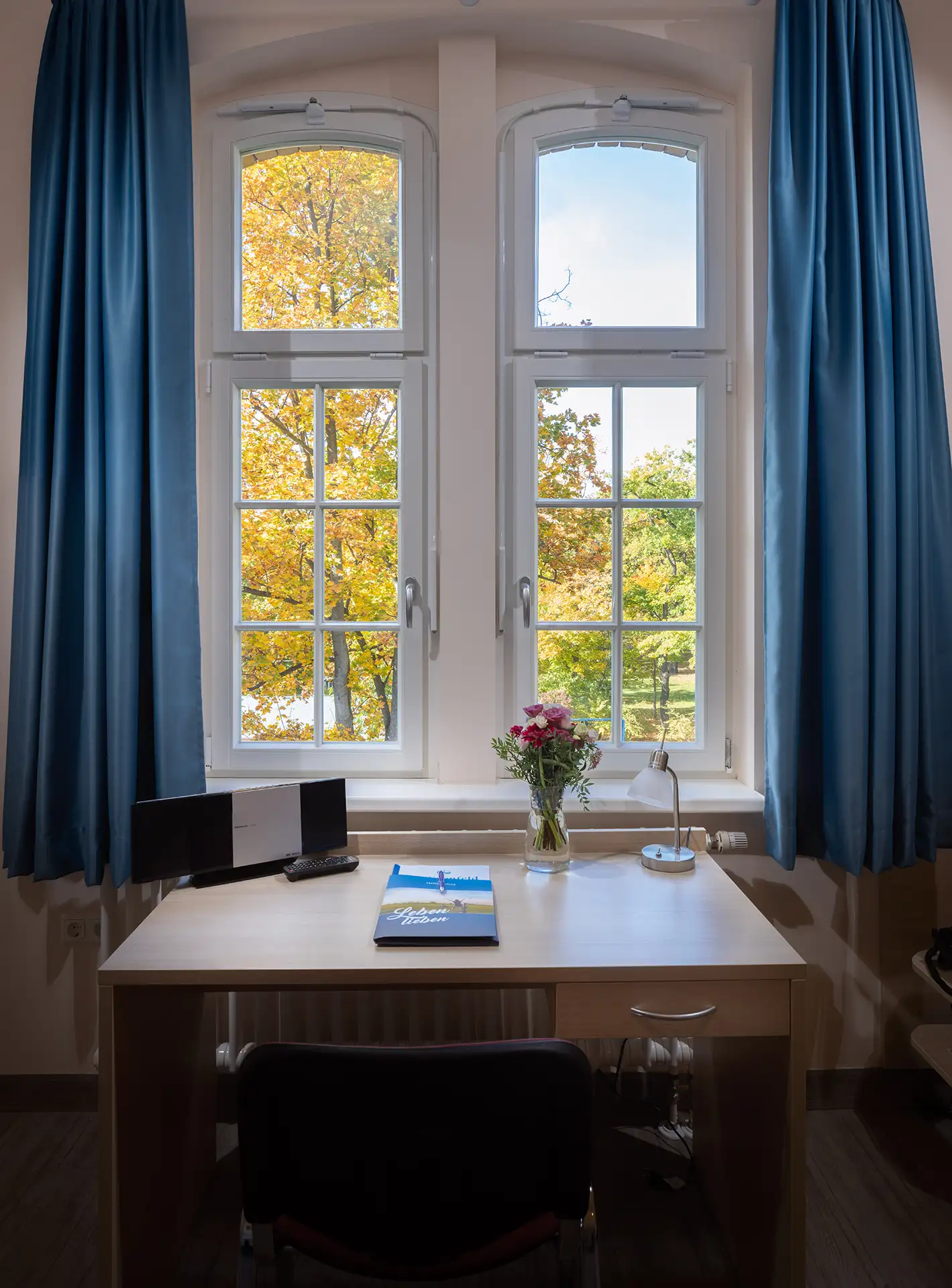 Bild. Beschränke dich dabei auf maximal 12 Wörter. Schreibtisch mit Blumen und Aussicht auf herbstliche Bäume durch Doppelfenster.