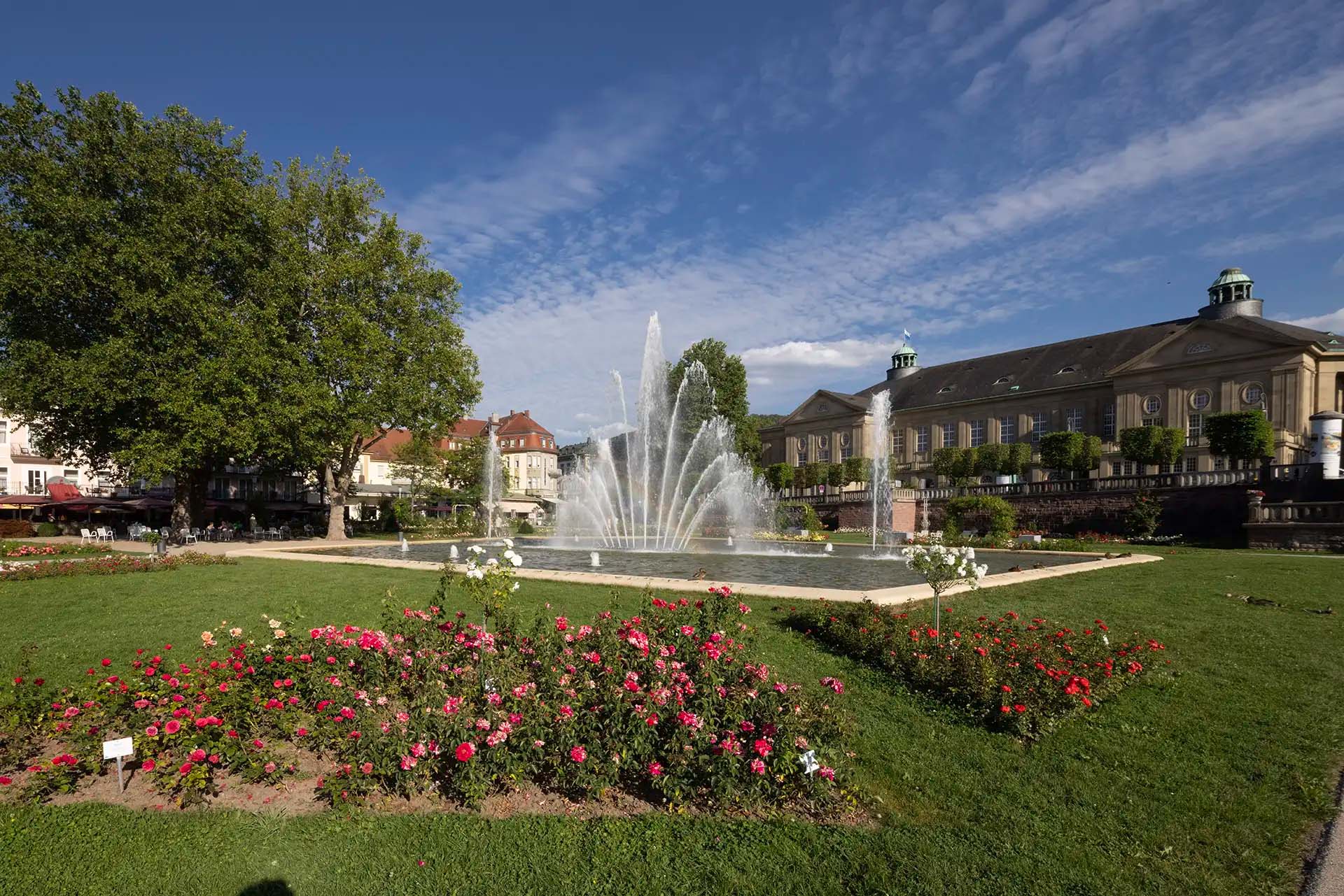 Springbrunnen und Blumenbeet im Park vor historischem Gebaeude.