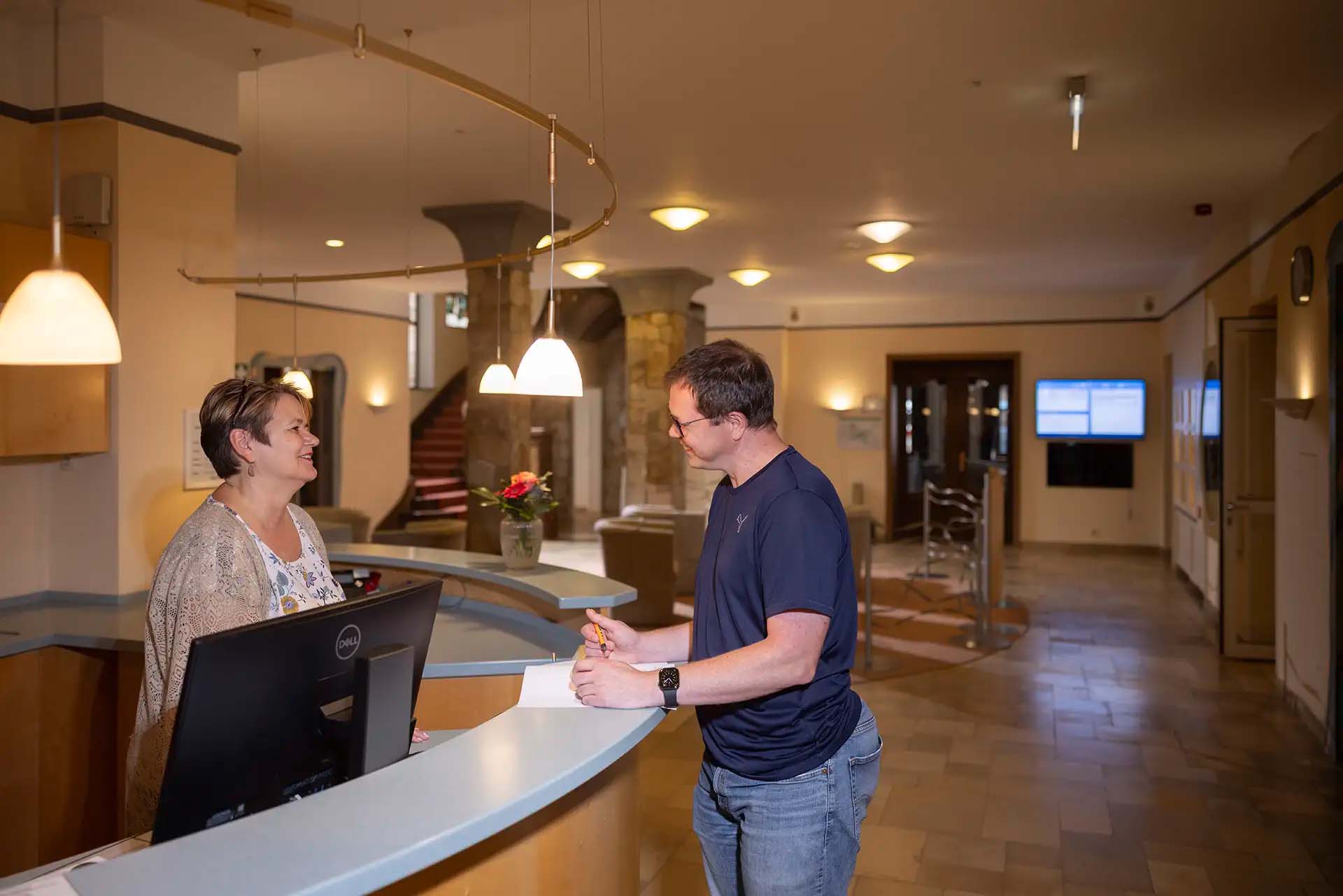 Mitarbeiter an Rezeption laechelt und spricht mit einem Gast.