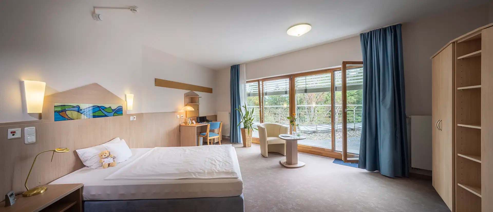 Helles Patientenzimmer mit Bett, Schreibtisch und Zugang zu Balkon.