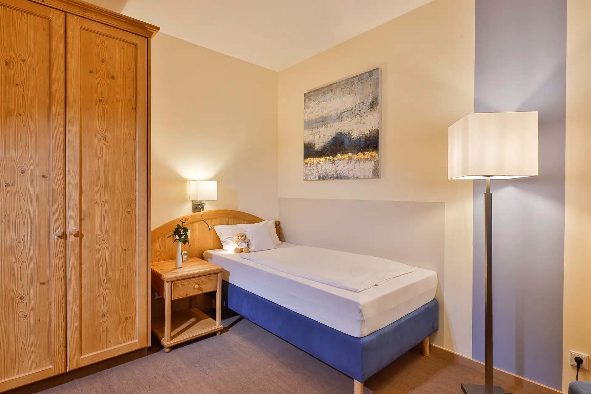 Gemütliches Patientenzimmer mit Einzelbett, Schrank und Nachttischlampe.