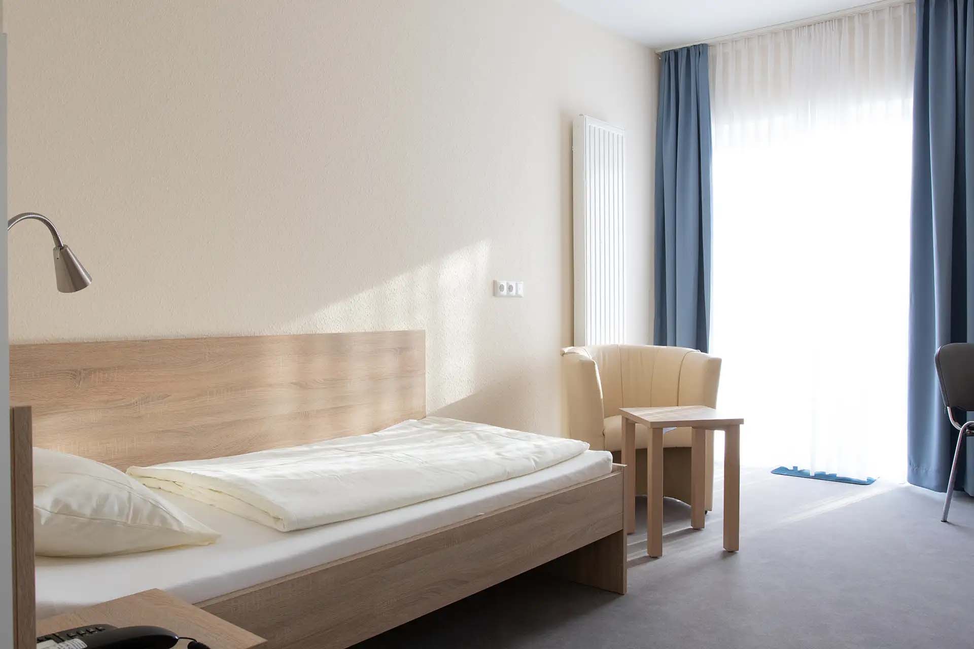 Einzelzimmer mit Bett, Sessel, Tisch und großen Fenstern.