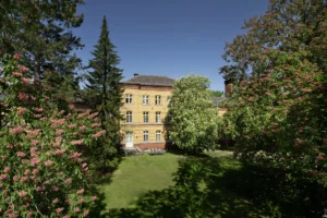 Gelbes Backsteingebäude mit grünen Bäumen und Personen im Innenhof.