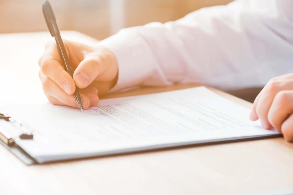 Hand schreibt auf ein Dokument mit Klemmbrett im Hintergrund.