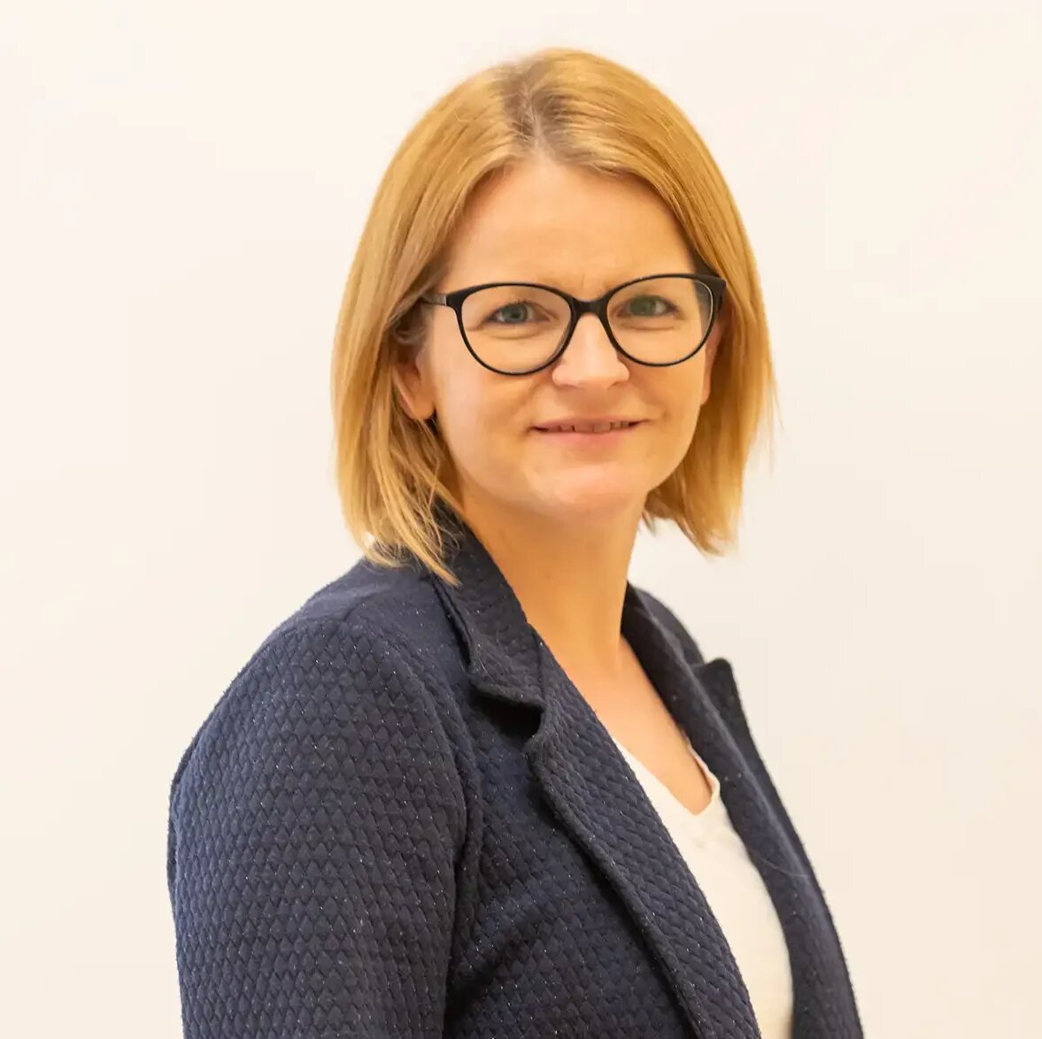 Porträt einer Frau mit Brille und dunklem Blazer vor hellem Hintergrund.