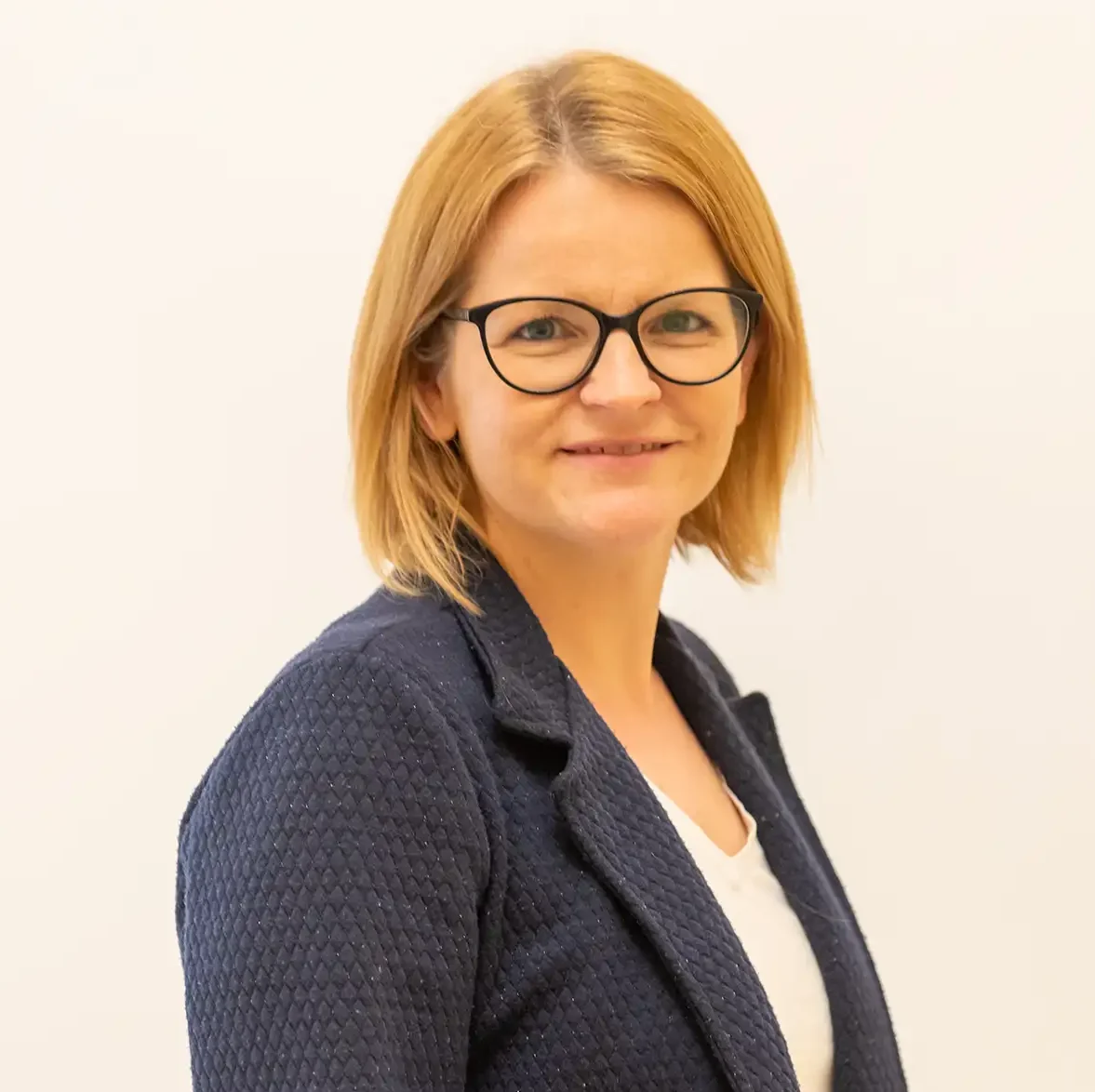 Porträt einer Frau mit Brille und dunklem Blazer vor hellem Hintergrund.