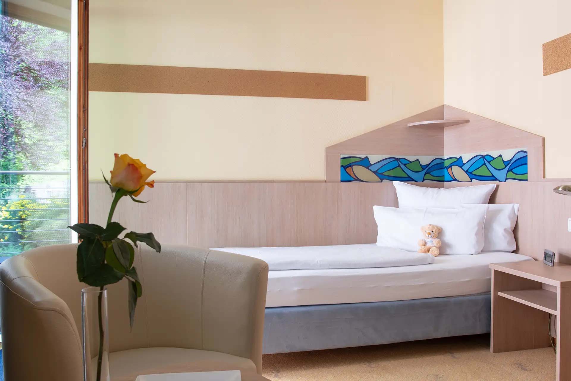 Gemütliches Patientenzimmer mit Einzelbett, Sessel, Teddybär und Rose