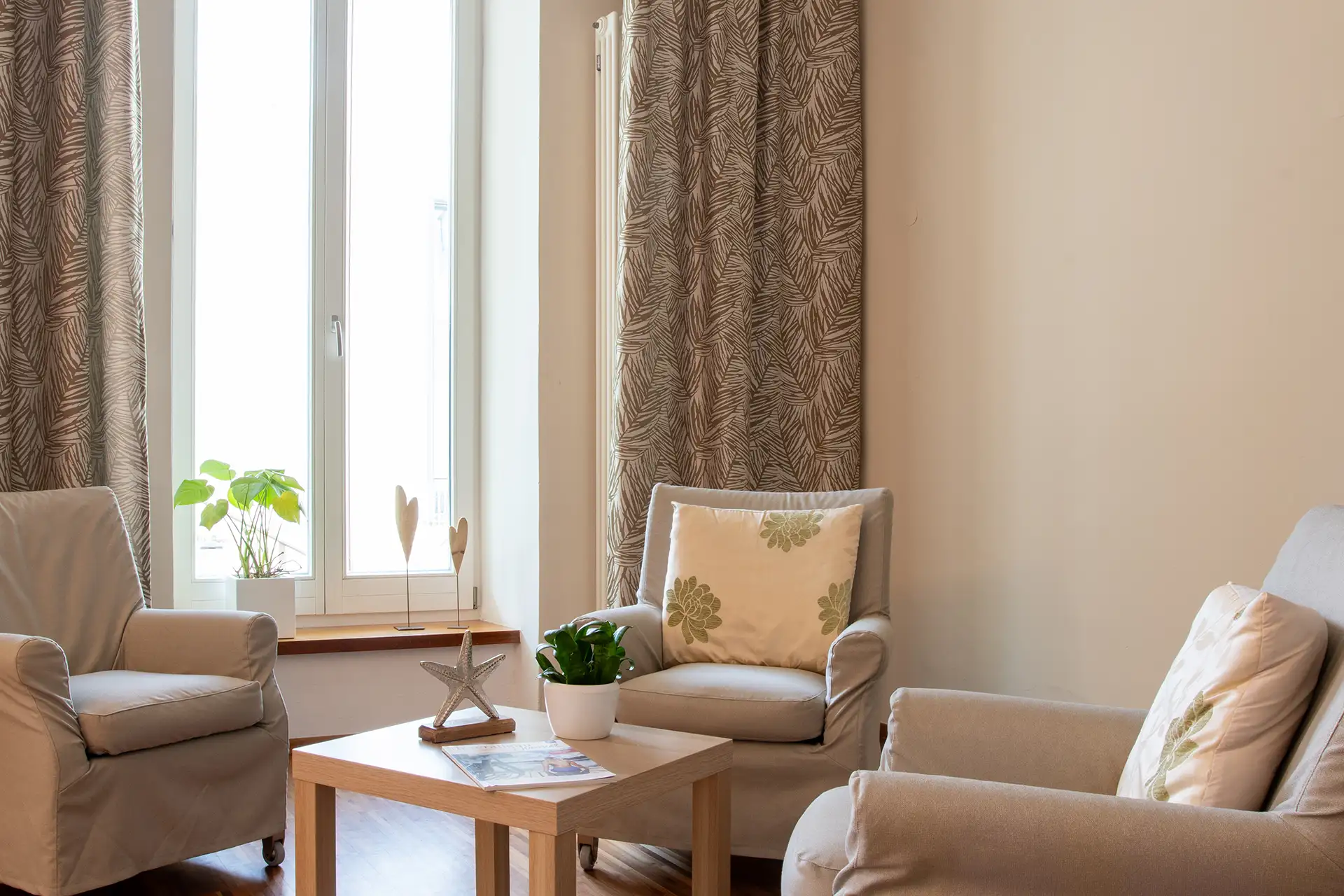 Gemütliche Sitzecke mit Sesseln, Kissen und Tischpflanzen vor großem Fenster.