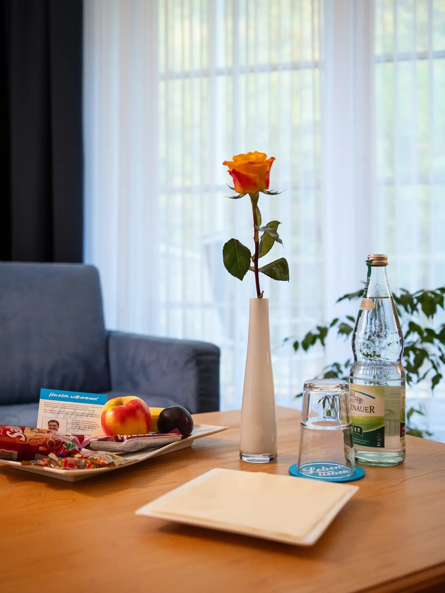 Willkommenstisch mit Rose, Obstteller, Getränk und Willkommenskarte in gemütlicher Umgebung.
