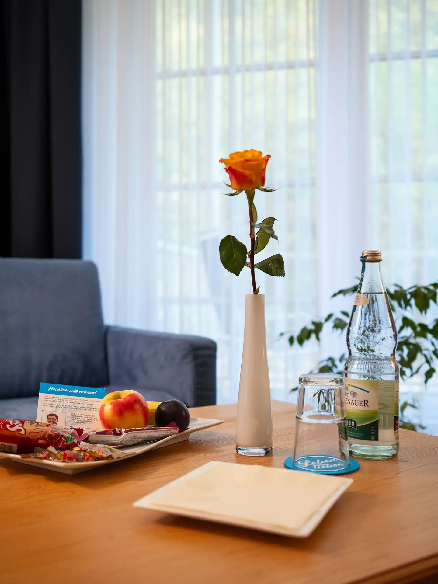 Willkommenstisch mit Rose, Obstteller, Getränk und Willkommenskarte in gemütlicher Umgebung.