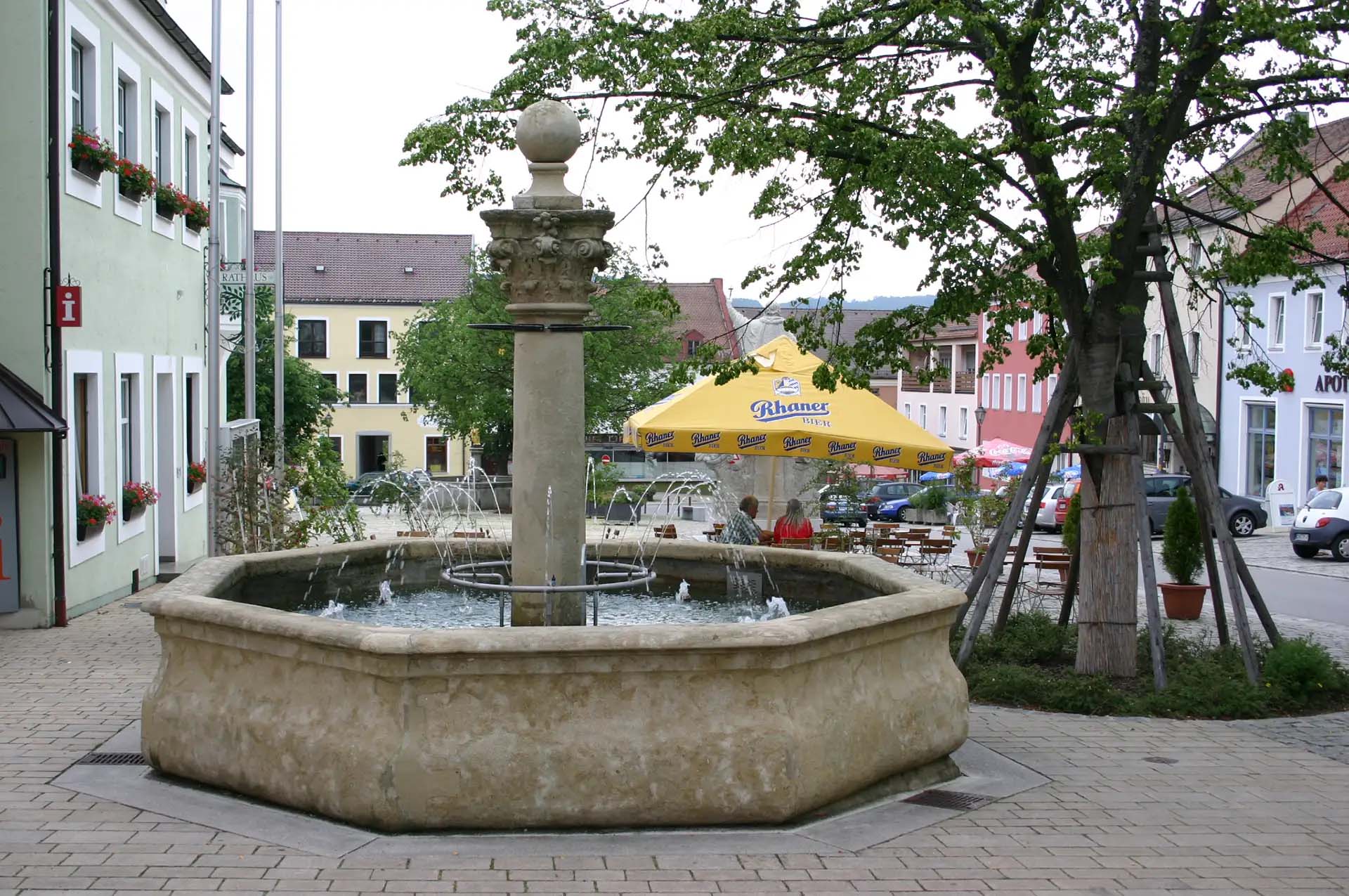 Brunnen auf einem Platz mit umliegenden Häusern und Biergarten-Schirm.