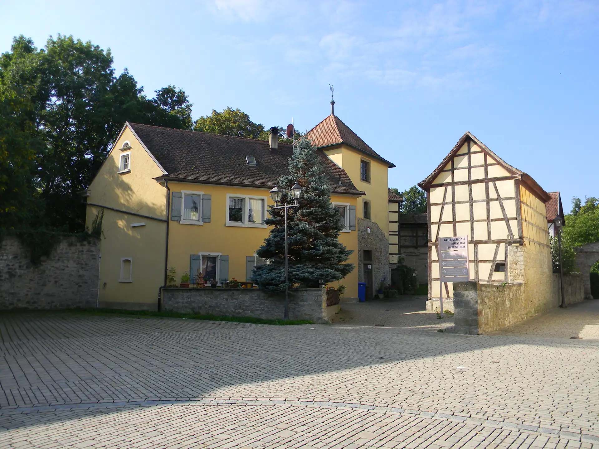 Historische Fachwerkhäuser an einem gepflasterten Platz, idyllisches Ambiente mit Bäumen und Pflanzen.