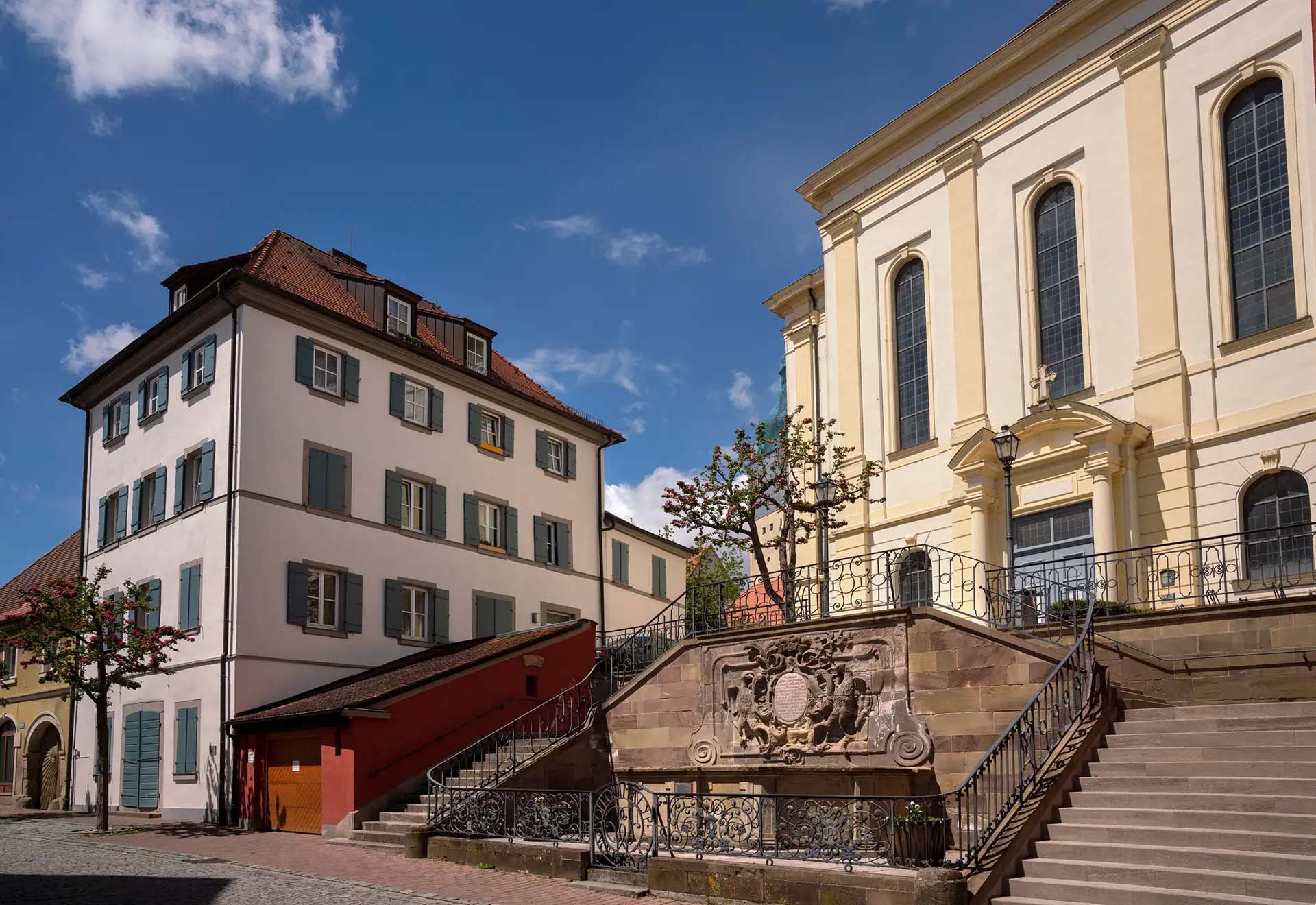 Historischer Platz mit Treppen, Steinwappen und Gebäuden unter blauem Himmel und Sonnenschein.