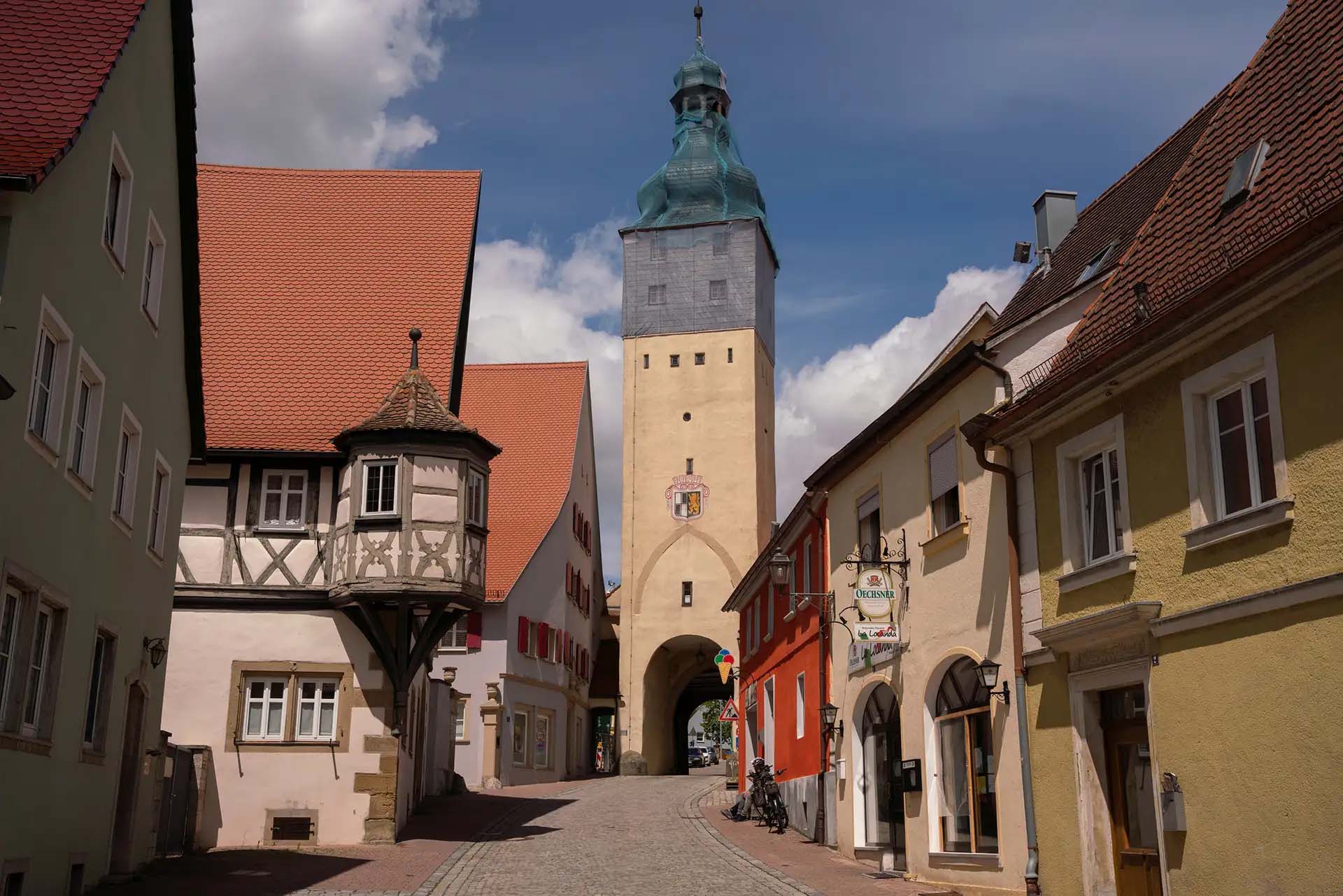 Historische Altstadtstraße mit Fachwerkhaus, Torbogen und farbenfrohen Gebäuden unter blauem Himmel.