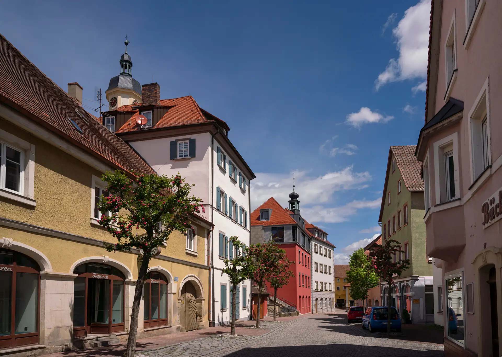 Altstadtgasse mit charmanten Gebäuden, Kopfsteinpflaster, blühenden Bäumen und historischem Flair unter blauem Himmel.