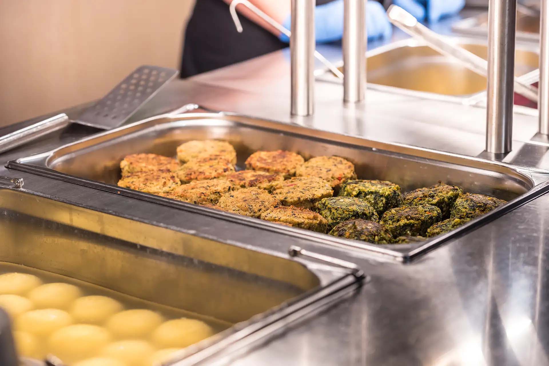 Eine Essensausgabe mit verschiedenen Frikadellen und Kartoffelklößen in Wärmebehältern.