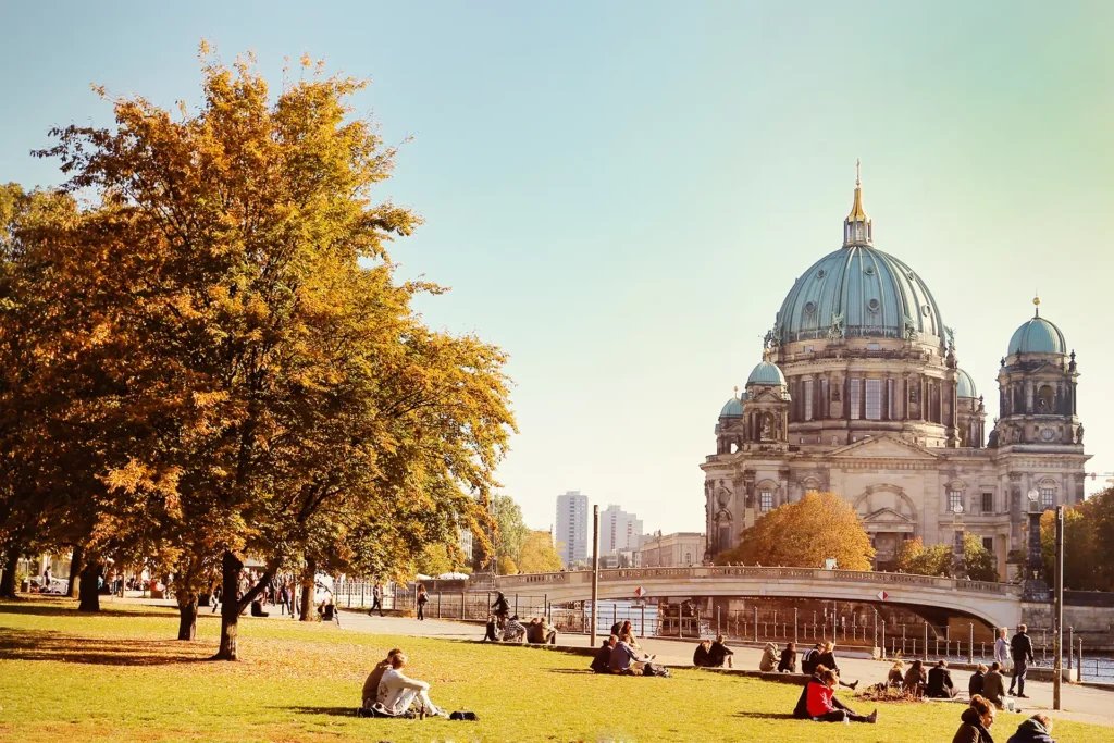 Herbstliche Szene in Berlin mit Blick auf den Berliner Dom. Menschen entspannen auf der Wiese in einem Park unter goldgelben Bäumen, während die Sonne einen warmen Glanz über die Umgebung wirft.
