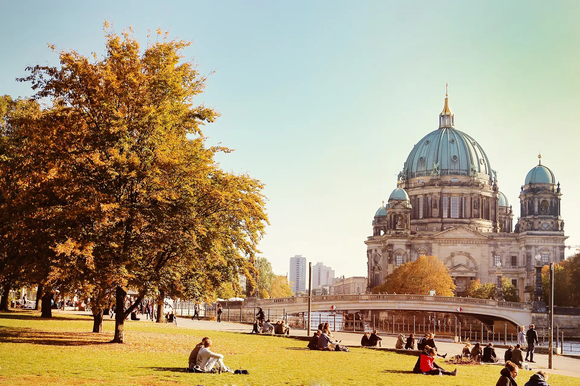 Herbstliche Szene in Berlin mit Blick auf den Berliner Dom. Menschen entspannen auf der Wiese in einem Park unter goldgelben Bäumen, während die Sonne einen warmen Glanz über die Umgebung wirft.