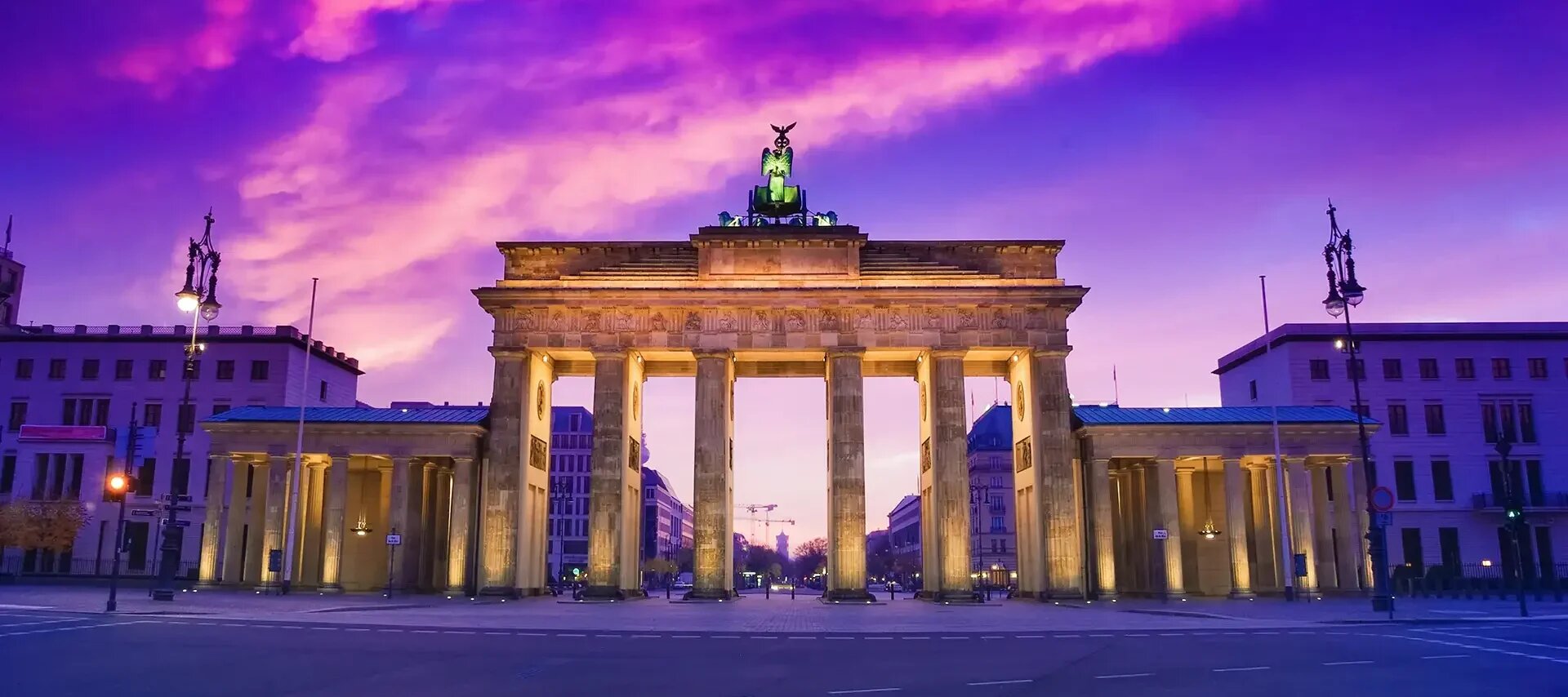 Das Brandenburger Tor in Berlin bei Sonnenuntergang. Der Himmel zeigt dramatische Violett- und Pinktöne, die das beleuchtete Tor eindrucksvoll hervorheben und eine magische Abendstimmung erzeugen.