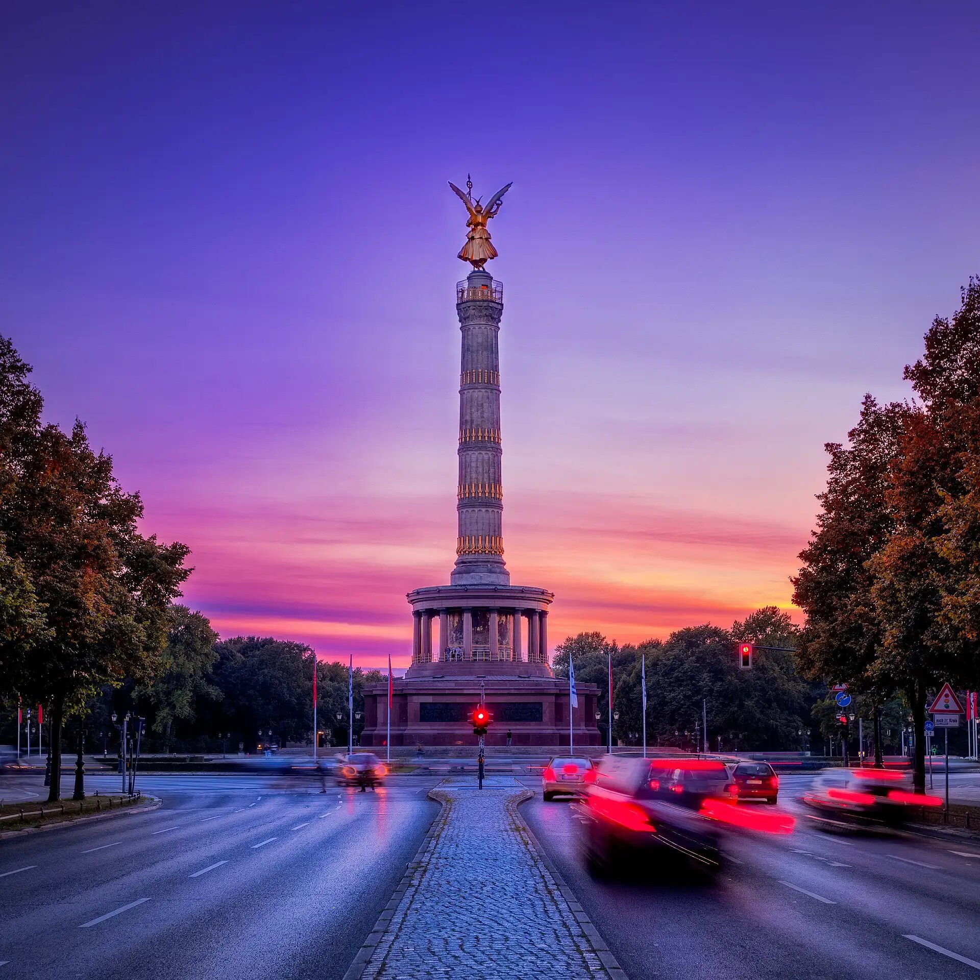 Die Berliner Siegessäule bei Sonnenuntergang, umgeben von lebendigem Verkehr und Bäumen.