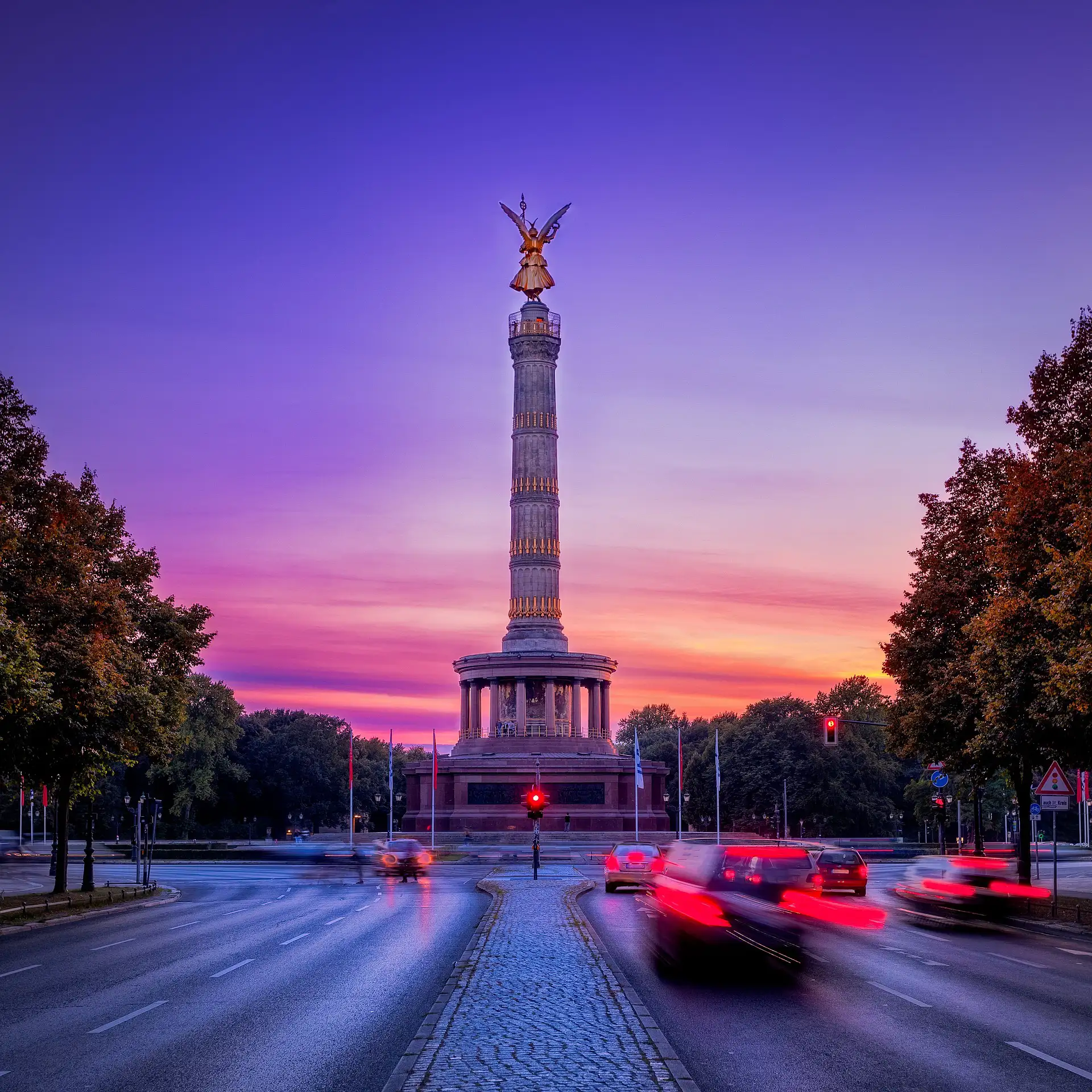 Die Berliner Siegessäule bei Sonnenuntergang, umgeben von lebendigem Verkehr und Bäumen.