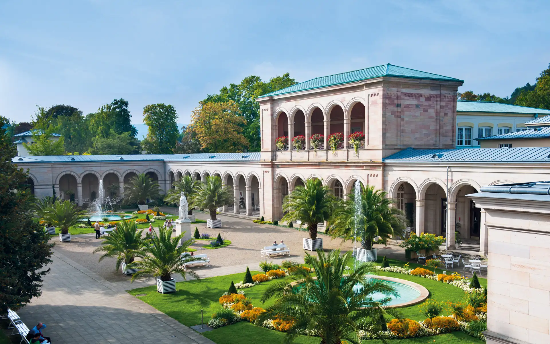 Historischer Kurgarten mit Palmen, Springbrunnen und blumengeschmücktem Arkadenbau.