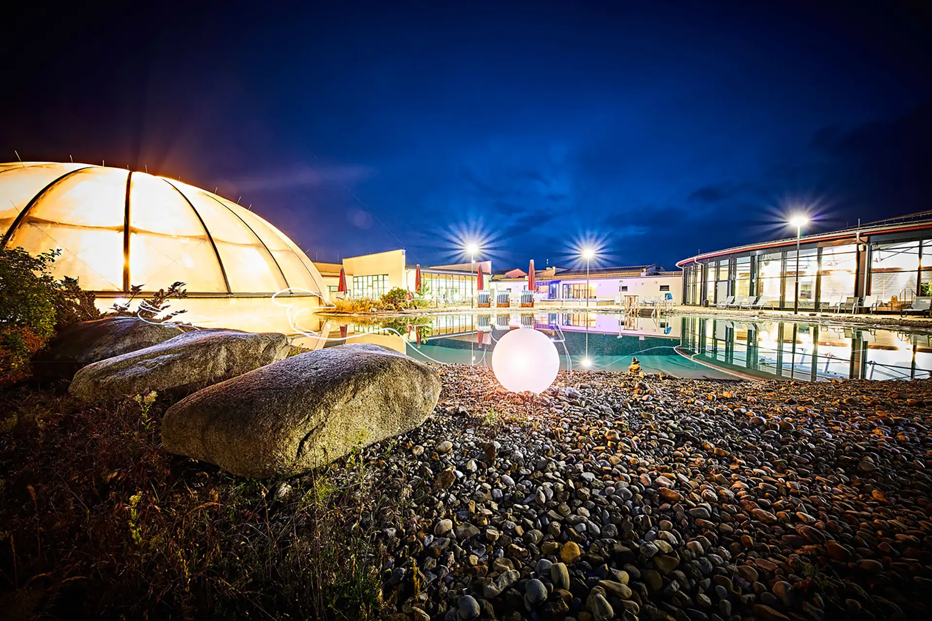 Thermalbad bei Nacht mit beleuchteter Kuppel, Außenpool und Steingarten.