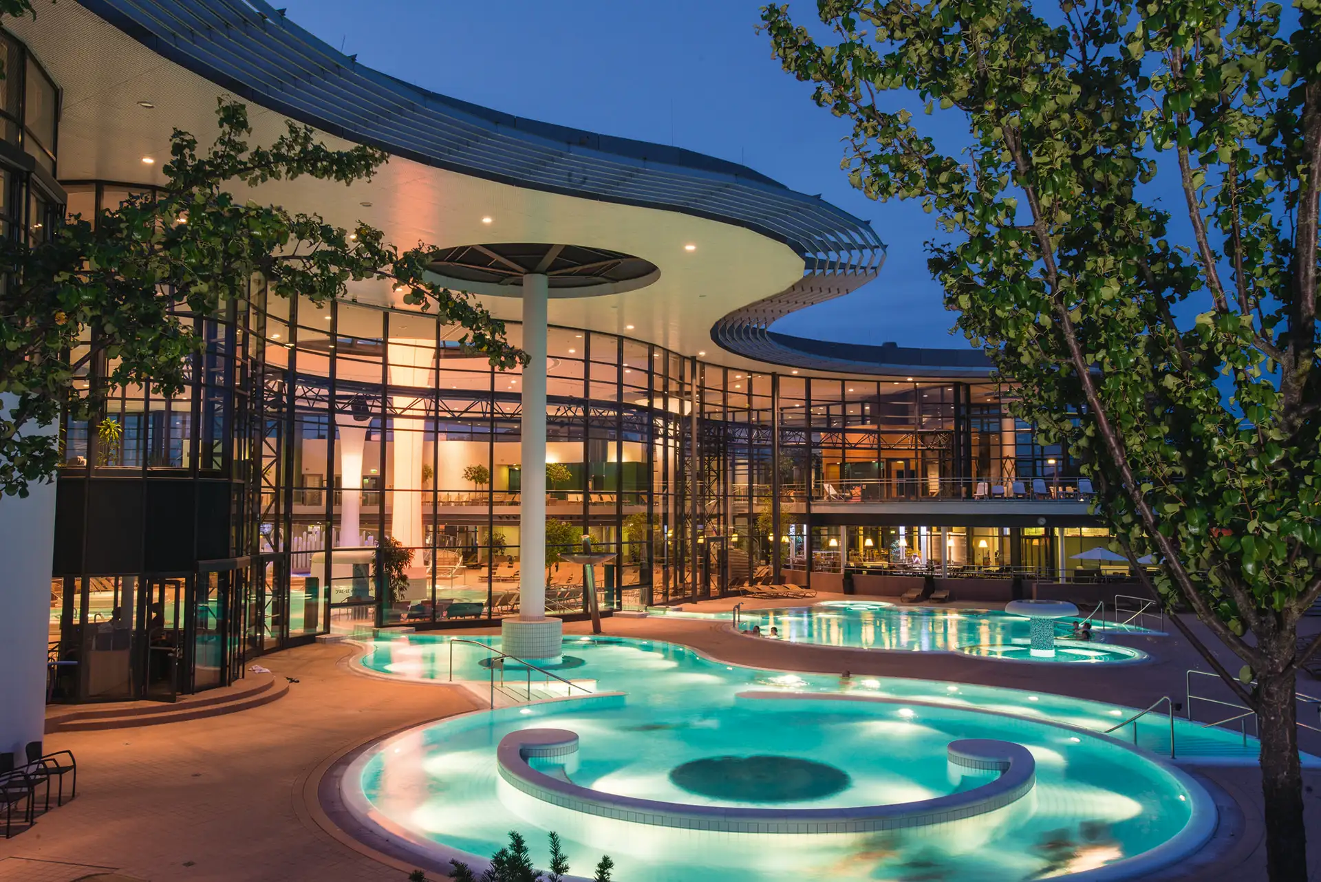 Modernes Thermalbad bei Nacht mit beleuchteten Außenpools und beeindruckender Glasarchitektur.